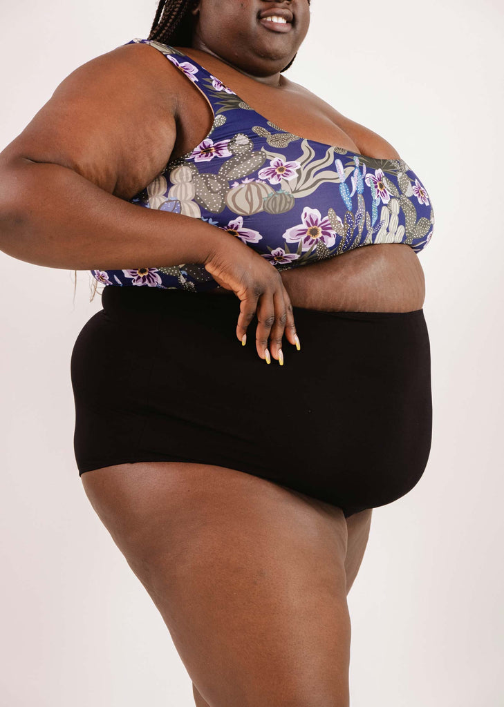 Person wearing a floral sports bra and Bermudes Black extra high waisted bikini bottom by Mimi & August, presenting a flattering silhouette against a white background.