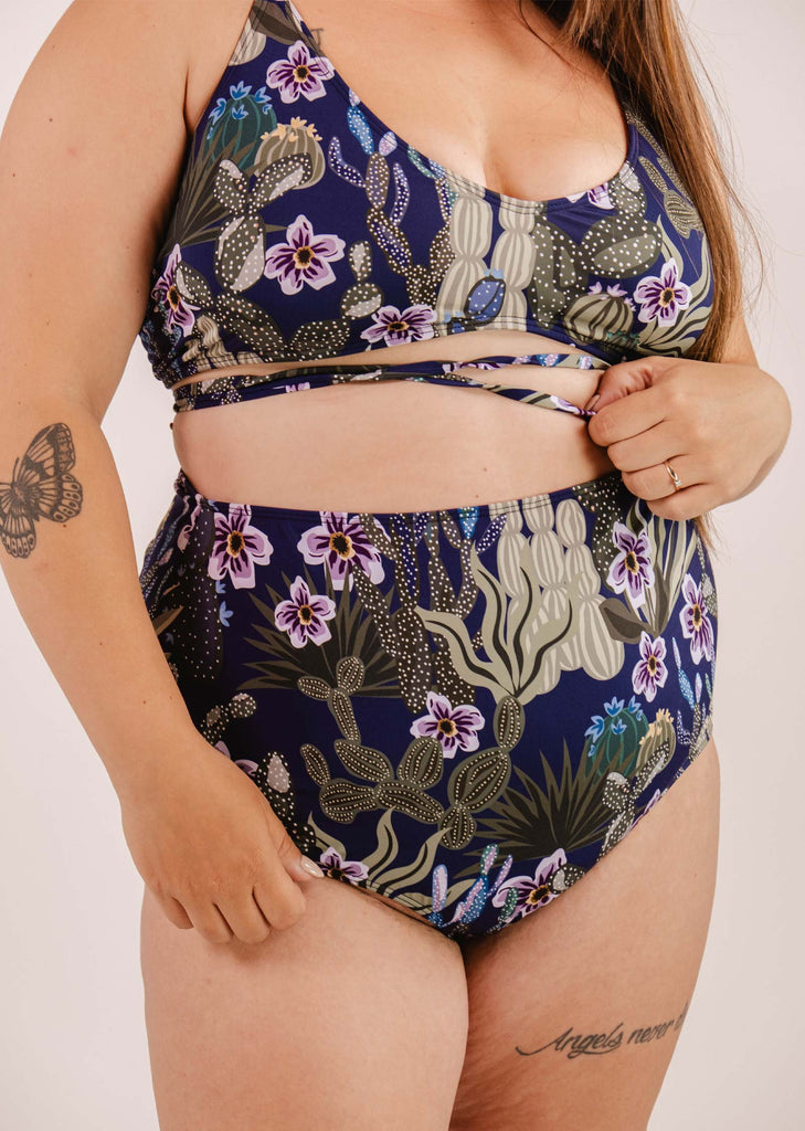 A person is wearing a blue floral-patterned swimsuit with a crossover strap design and a Bermudes Jardin de Nuit extra high waisted bikini bottom by Mimi & August. The person's tattoo of a butterfly on their arm and a script tattoo on their thigh are visible.