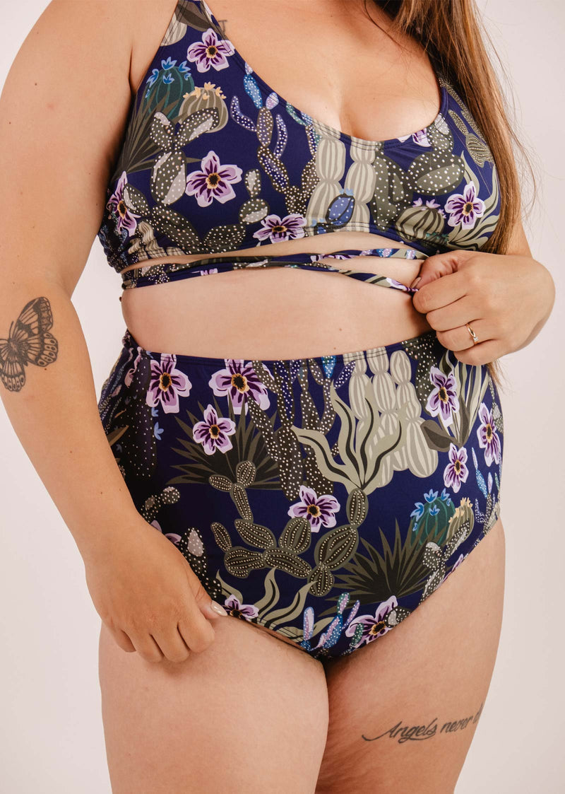 Une personne porte un maillot de bain bleu à motifs floraux avec des bretelles croisées et un bas de bikini à taille très haute Bermudes Jardin de Nuit de Mimi & August. Le tatouage d'un papillon sur le bras et un tatouage d'écriture sur la cuisse sont visibles.