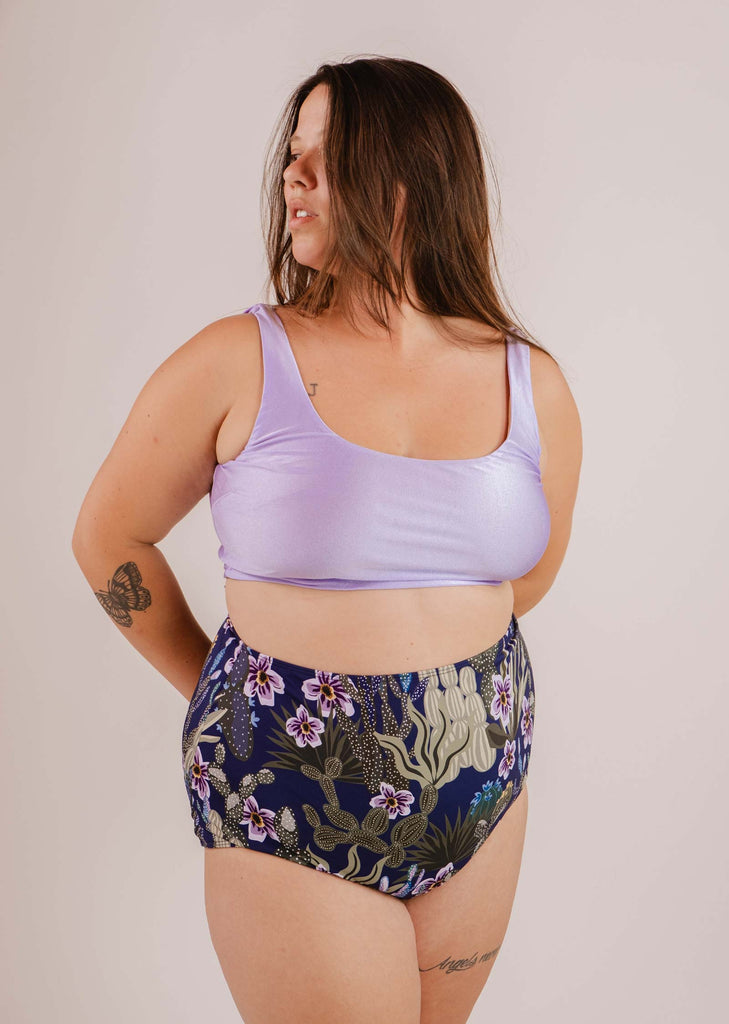 Une personne aux cheveux longs vêtue d'un haut sans manches violet et d'un bas de bikini à taille extra haute Bermudes Jardin de Nuit de Mimi & August, debout sur un fond uni. Elle a des tatouages visibles sur le bras gauche et la hanche.