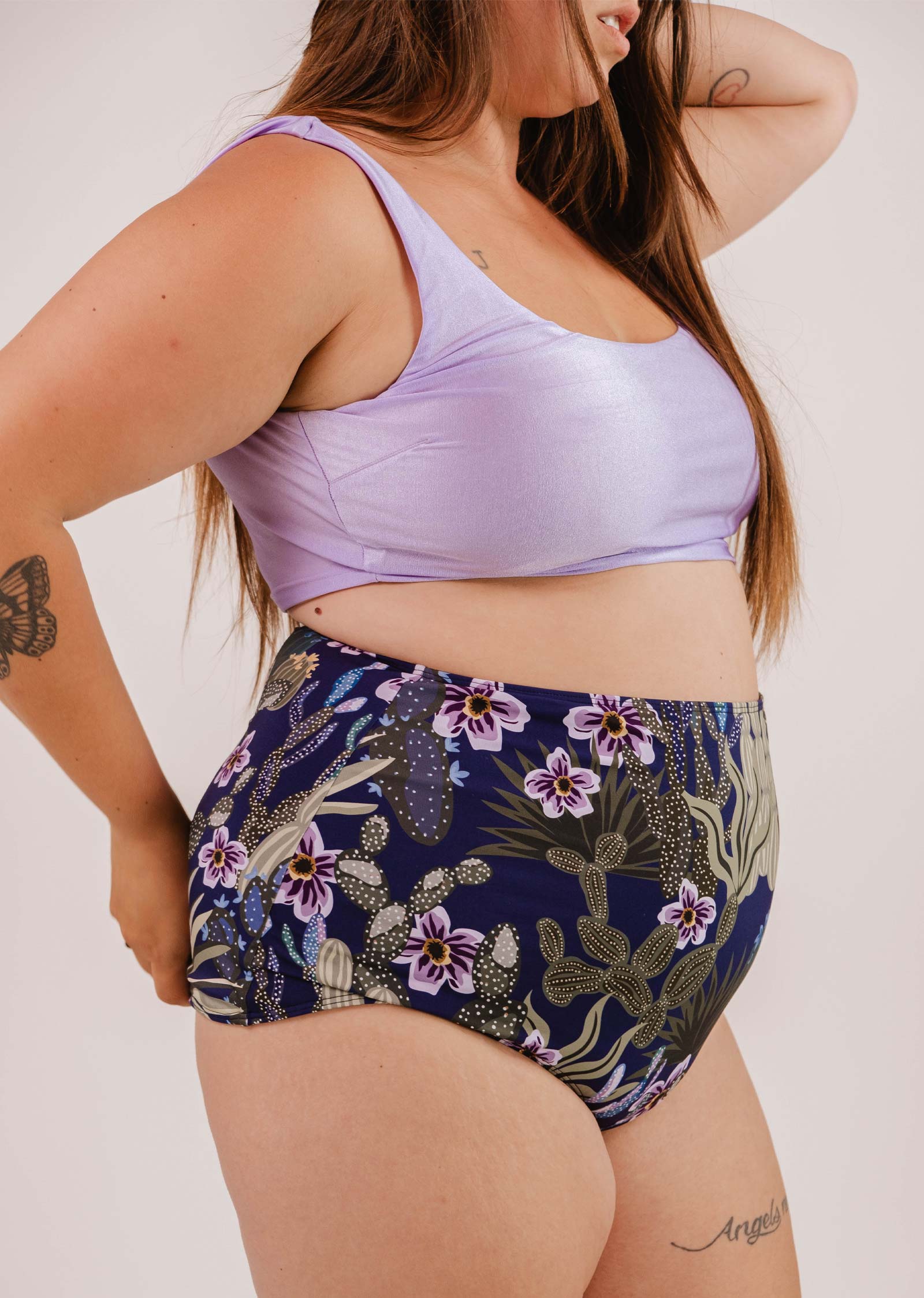 A person poses in a lilac crop top and Mimi & August Bermudes Jardin de Nuit extra high waisted bikini bottom. They have long hair and tattoos on their arm and leg.