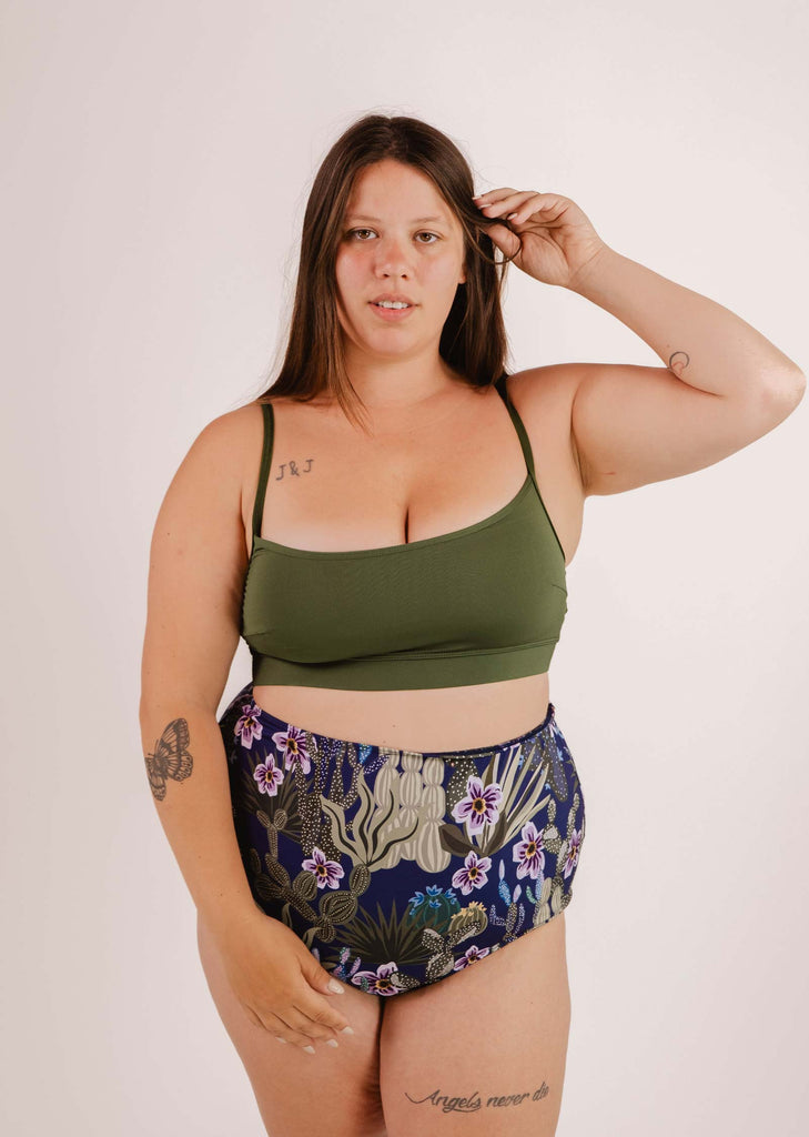 A woman with long brown hair wearing a green sports bra and Mimi & August Bermudes Jardin de Nuit extra high waisted bikini bottom stands against a plain background, holding a strand of her hair with her right hand.