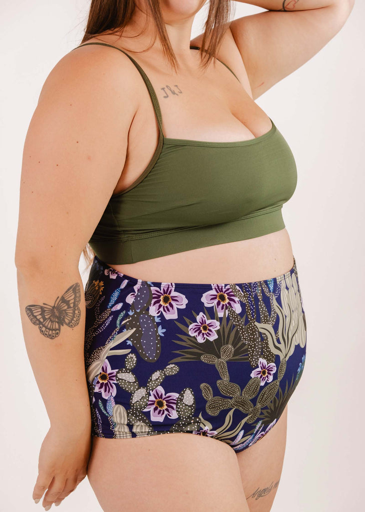 A person wearing a green top and Bermudes Jardin de Nuit extra high-waisted bikini bottom by Mimi & August with a floral and cactus print stands against a white background. Tattoos are visible on their arm and upper chest.