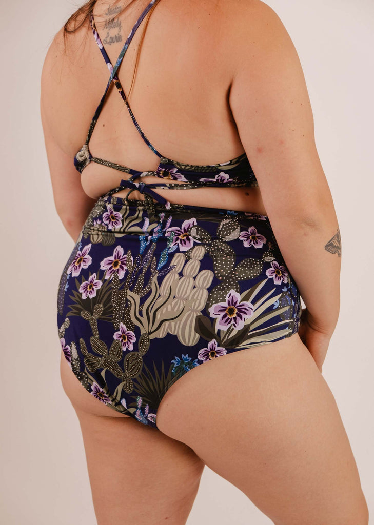 A person in a floral and cactus print swimsuit is shown from the back, displaying cross straps and various tattoos, complemented by a Bermudes Jardin de Nuit extra high waisted bikini bottom from Mimi & August.