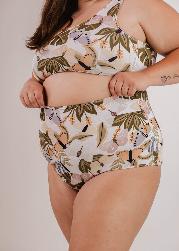 A person wearing a Mimi & August Bermudes Mariposa extra high waisted bikini bottom with a matching floral-patterned top adjusts the waistband, revealing a tattoo on their right forearm. The outfit is perfect for summer activities, showcasing the blend of style and comfort ideal for sunny days.