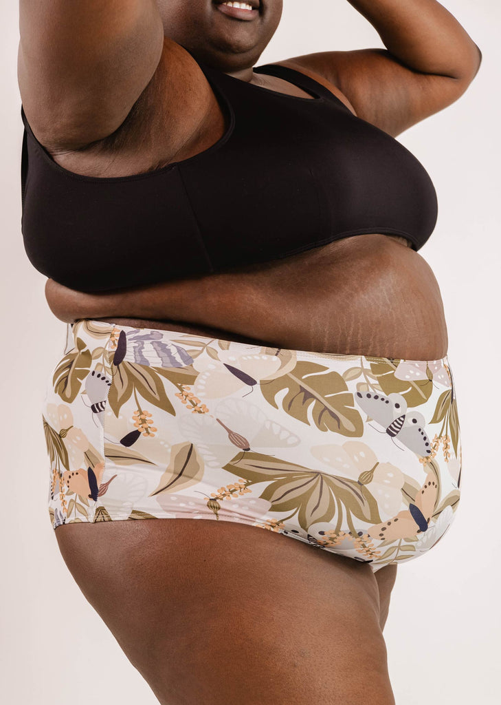 Person wearing a black top and patterned shorts with a leaf, animal, and butterfly print, raising their arms to showcase their flattering silhouette in the Bermudes Mariposa extra high waisted bikini bottom by Mimi & August.