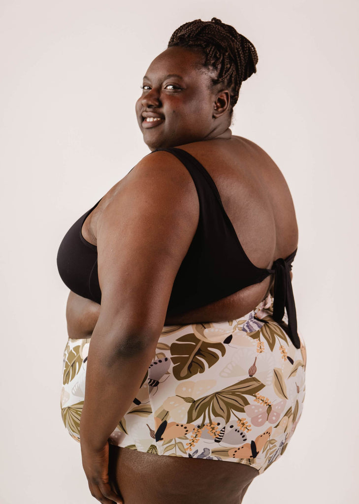 A person with braided hair is smiling while wearing a black and butterfly print two-piece swimsuit with a Bermudes Mariposa extra high waisted bikini bottom by Mimi & August, creating a flattering silhouette against a plain background.