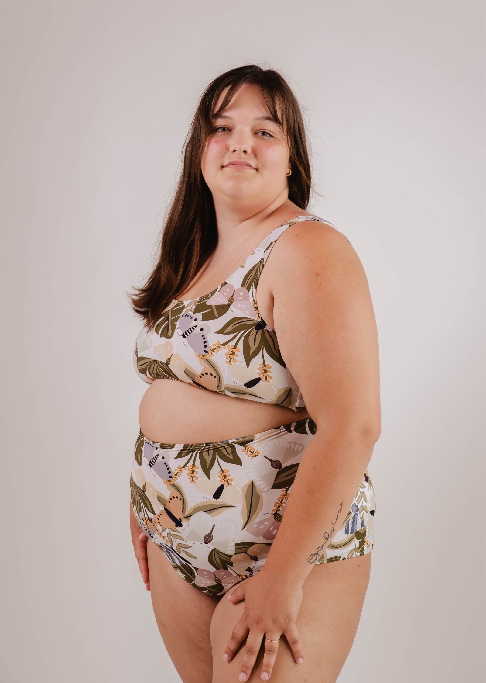 A person stands wearing a Mimi & August Bermudes Mariposa extra high waisted bikini bottom against a plain background. The individual, with shoulder-length brown hair and a relaxed expression, seems ready for summer activities.