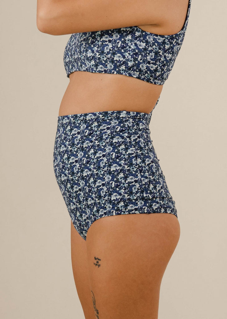 Une femme vêtue d'un haut de bikini bleu à fleurs et d'un bas de bikini Bermudes Moonflower extra taille haute