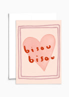 Cette carte de Saint-Valentin de Mimi & August, la carte de vœux Bisou Bisou, représente un cœur rose avec des lettres rouges sur du papier recyclé et est élégamment encadrée par des bordures à double ligne.