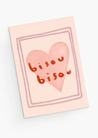 The Bisou Bisou Greeting Card from Mimi & August is made of recycled paper, showcasing a pink heart with "bisou bisou" in orange script at the center. Its simple border enhances its charm, making it ideal for sending love on Valentine's Day.