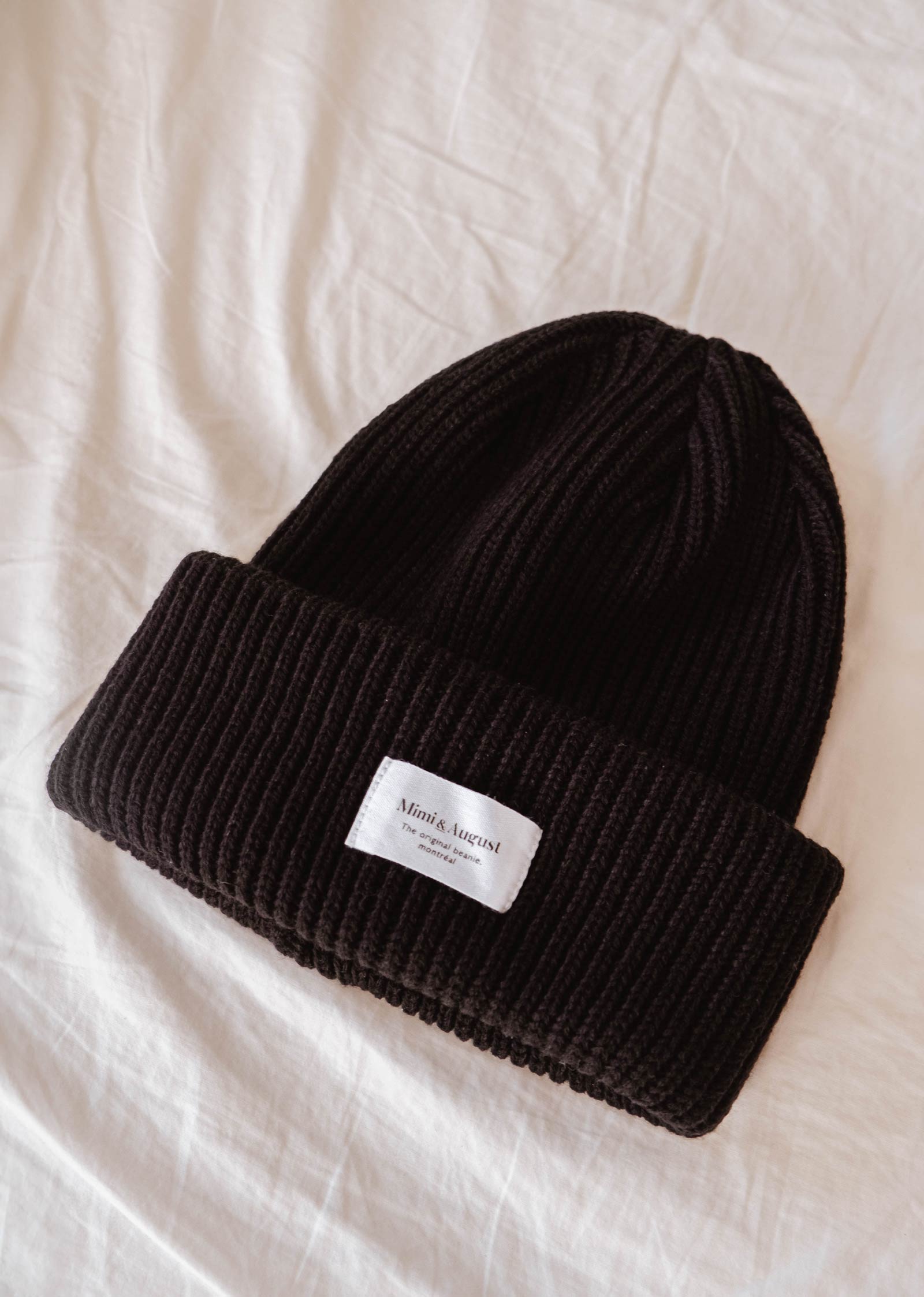 A Black Snuggle Beanie from Mimi & August, adorned with a small white label, lies on a white fabric surface, making it the perfect winter accessory.