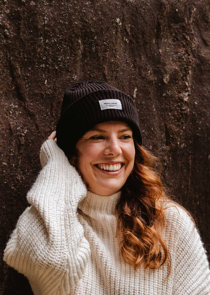 A woman with long hair is smiling while wearing a cream sweater and a cozy Black Snuggle Beanie by Mimi & August, standing in front of a textured wall.