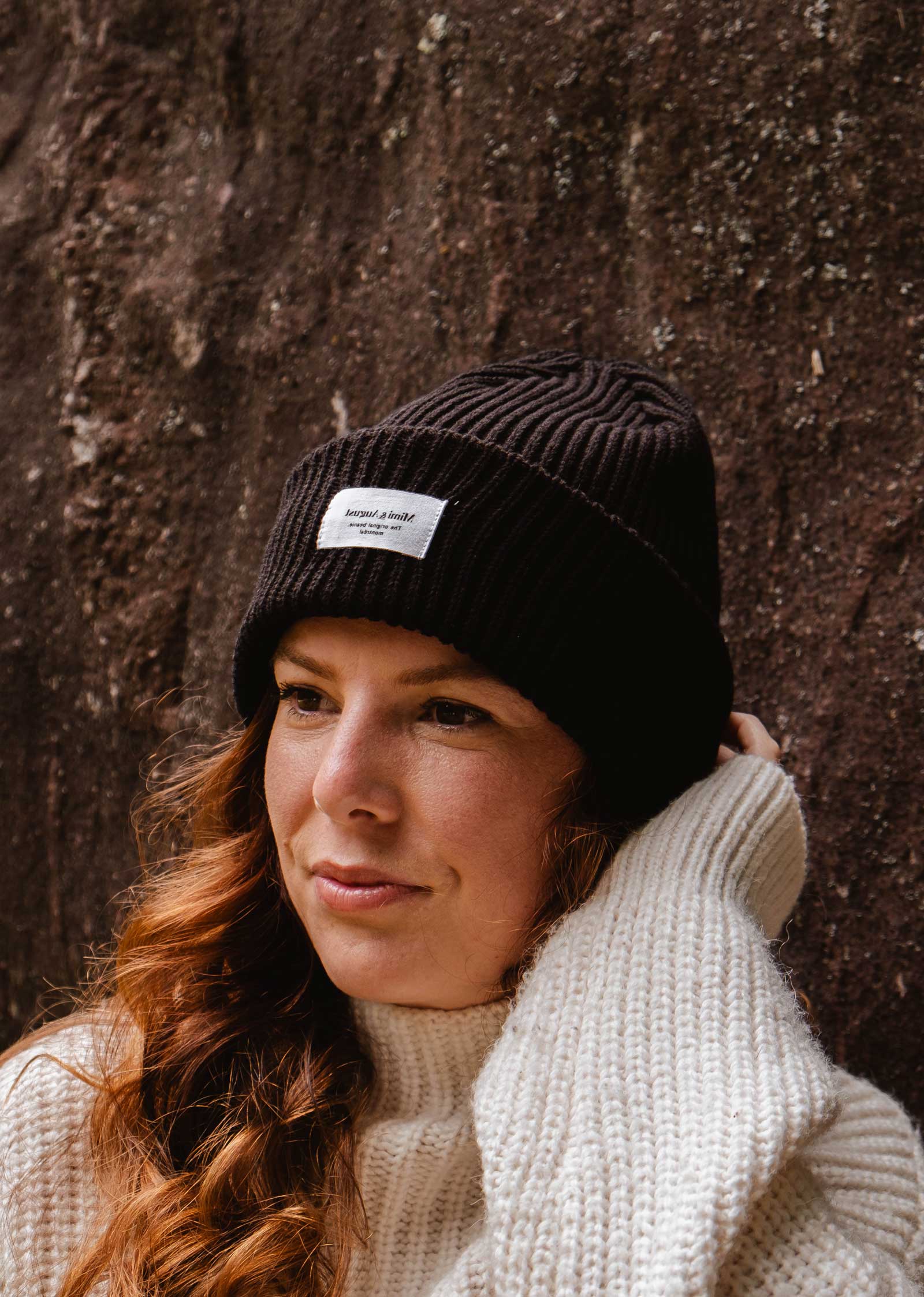 A person with long wavy hair, wearing a Black Snuggle Beanie by Mimi & August and a white sweater, stands against a rough-textured wall.