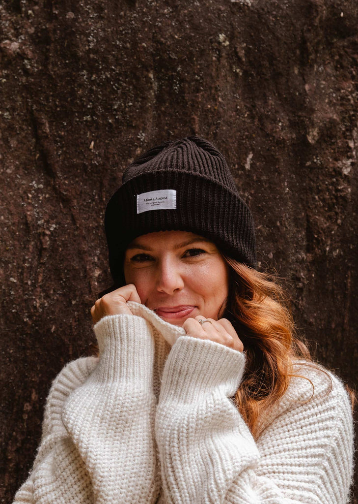 A person with long hair wears a Black Snuggle Beanie from Mimi & August and a white knit sweater, standing in front of a textured dark background, smiling and holding the sweater collar close to their face.