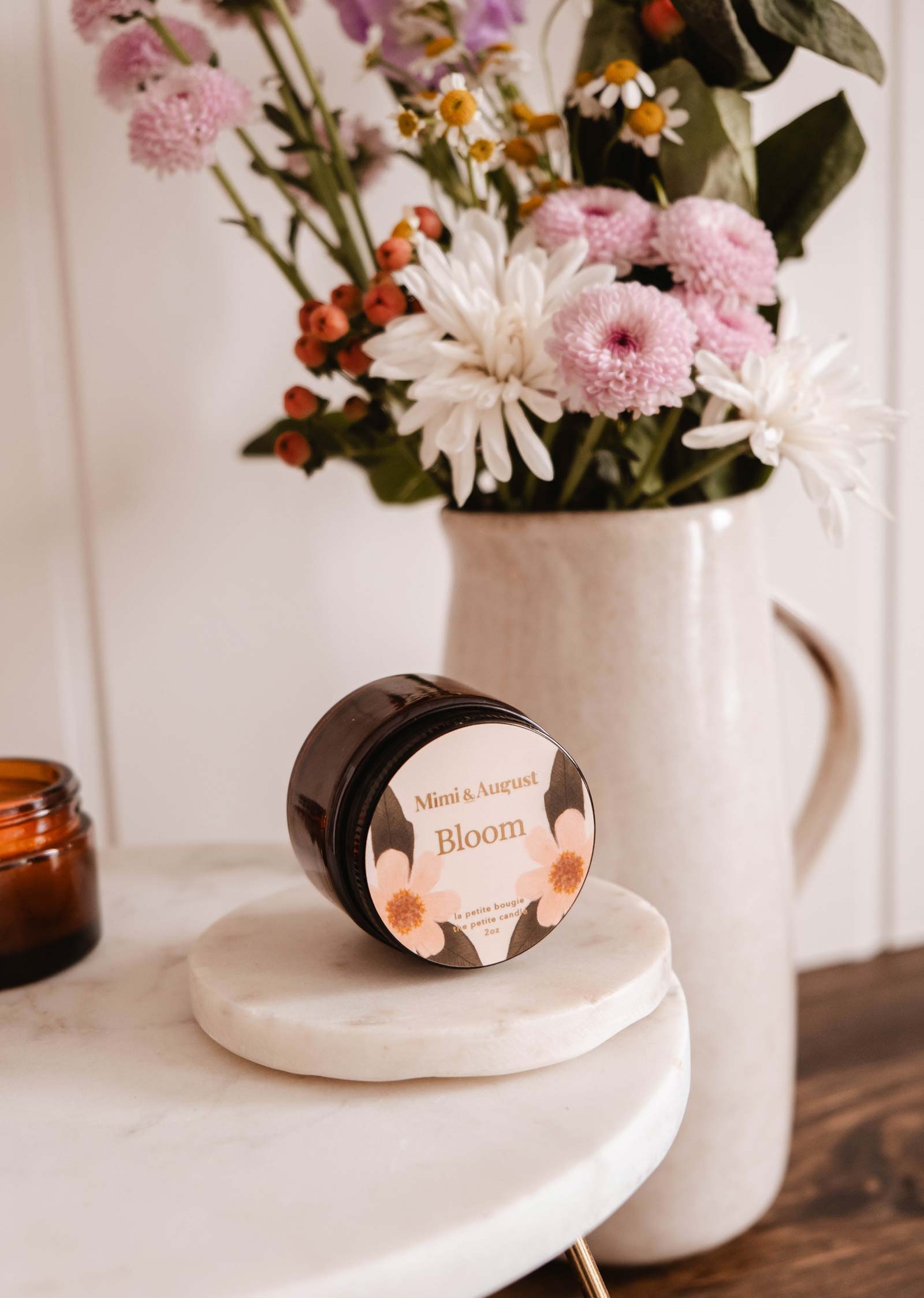 A round marble slab holds a "Mimi & August Bloom - Reusable Candle" jar, placed before a beige vase filled with pink, white, and orange flowers, capturing the candle's fruity essence.