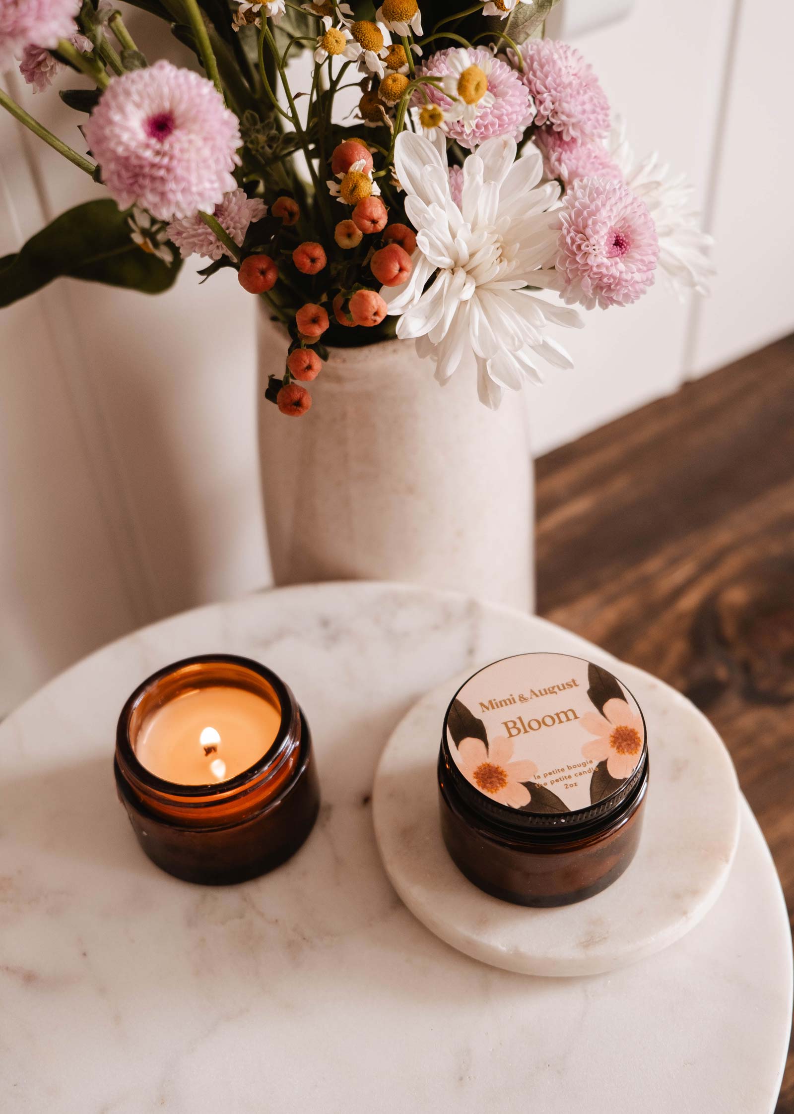 Une bougie fruitée allumée Mimi & August Bloom dans un pot étiqueté trône élégamment sur une table ronde en marbre, avec un vase de fleurs roses, blanches et orange complétant la scène.