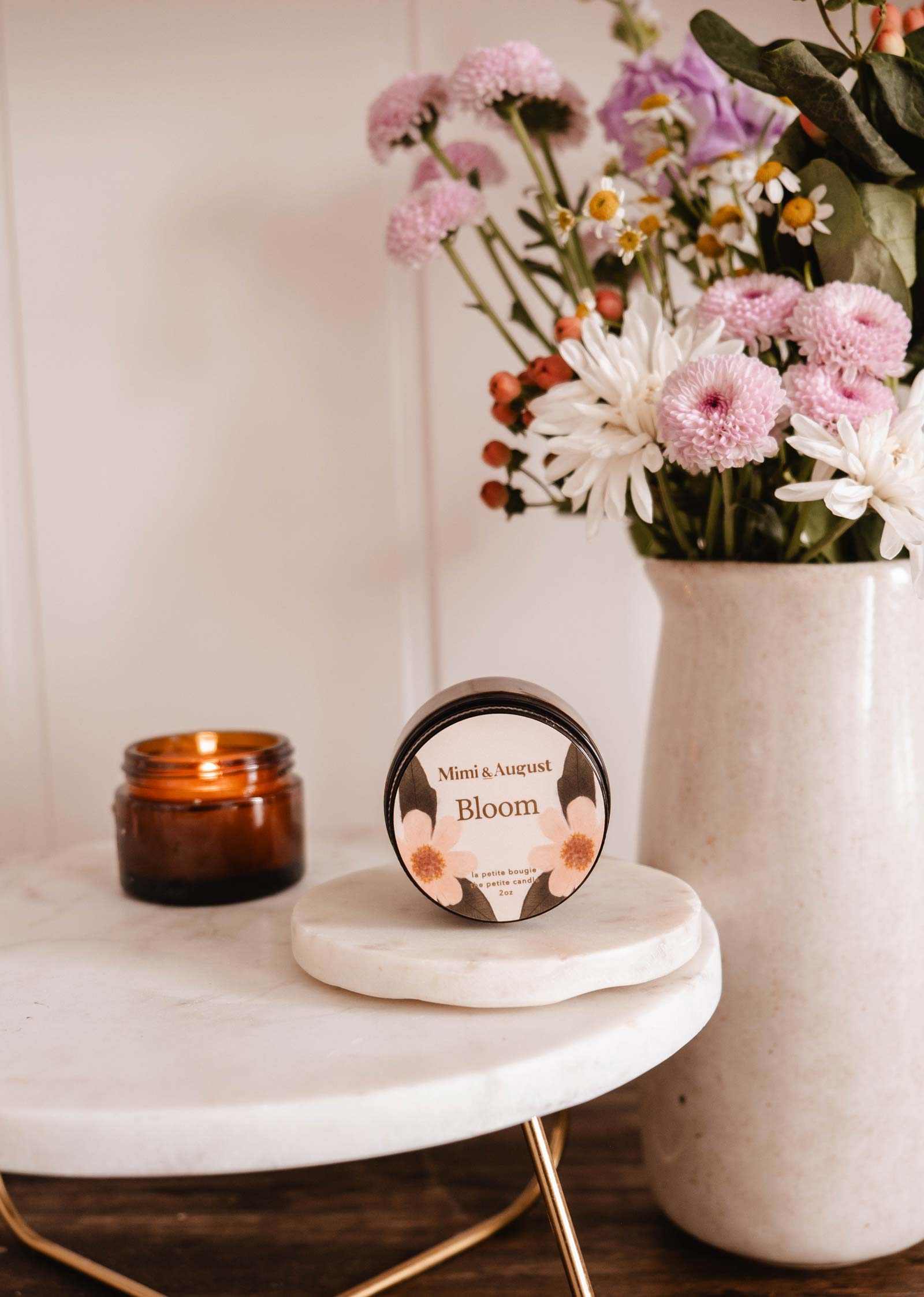 Une bougie de soya réutilisable Bloom de Mimi & August repose sur une surface en marbre blanc à côté d'un pot de bougie brun allumé et d'un vase de fleurs roses et blanches.