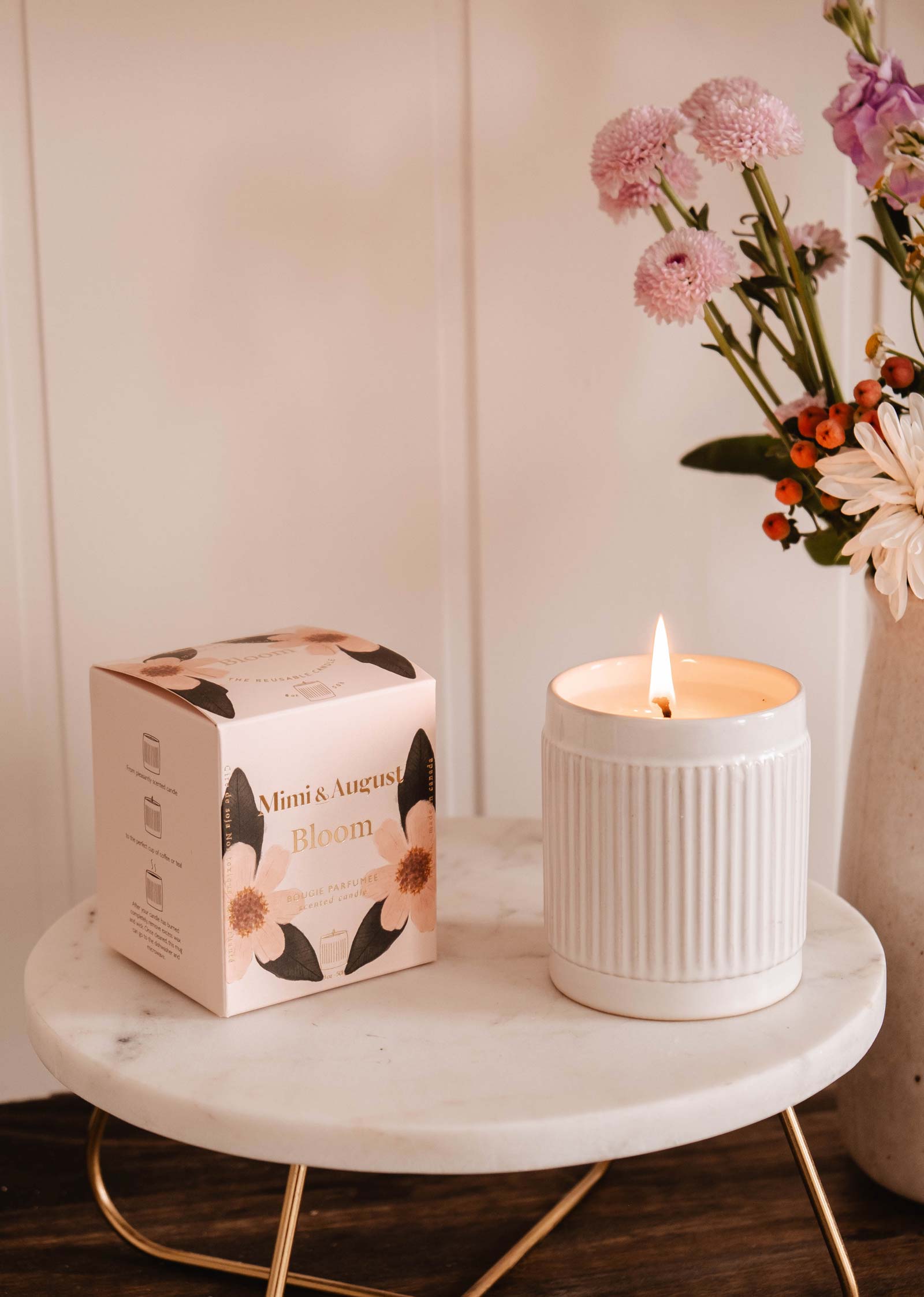 A lit Mimi & August Bloom candle in a ribbed ceramic jar sits on a round marble table beside its box, casting a warm glow. In the background, vibrant pink and orange flowers burst with fruity energy, creating an inviting atmosphere.