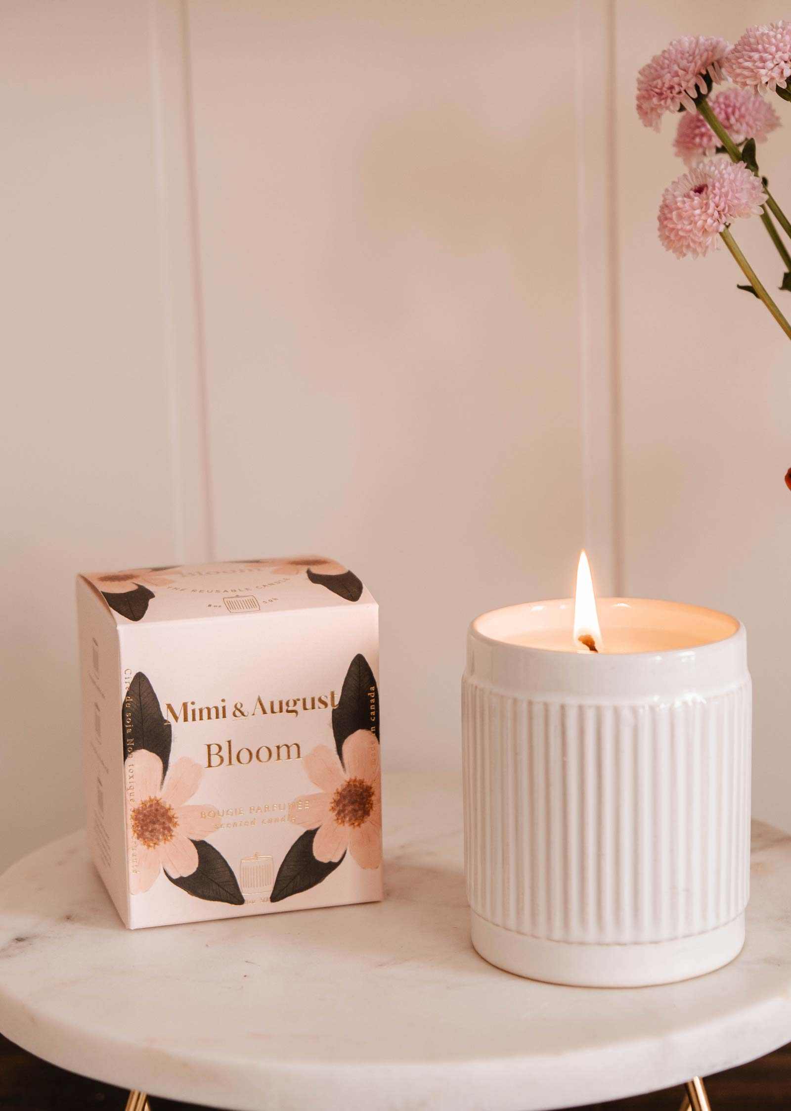 A lit Mimi & August Bloom – Reusable Soy Candle with an orange cranberry scent sits on a marble surface beside a floral "Bloom" box, with pink flowers in a vase in the background.