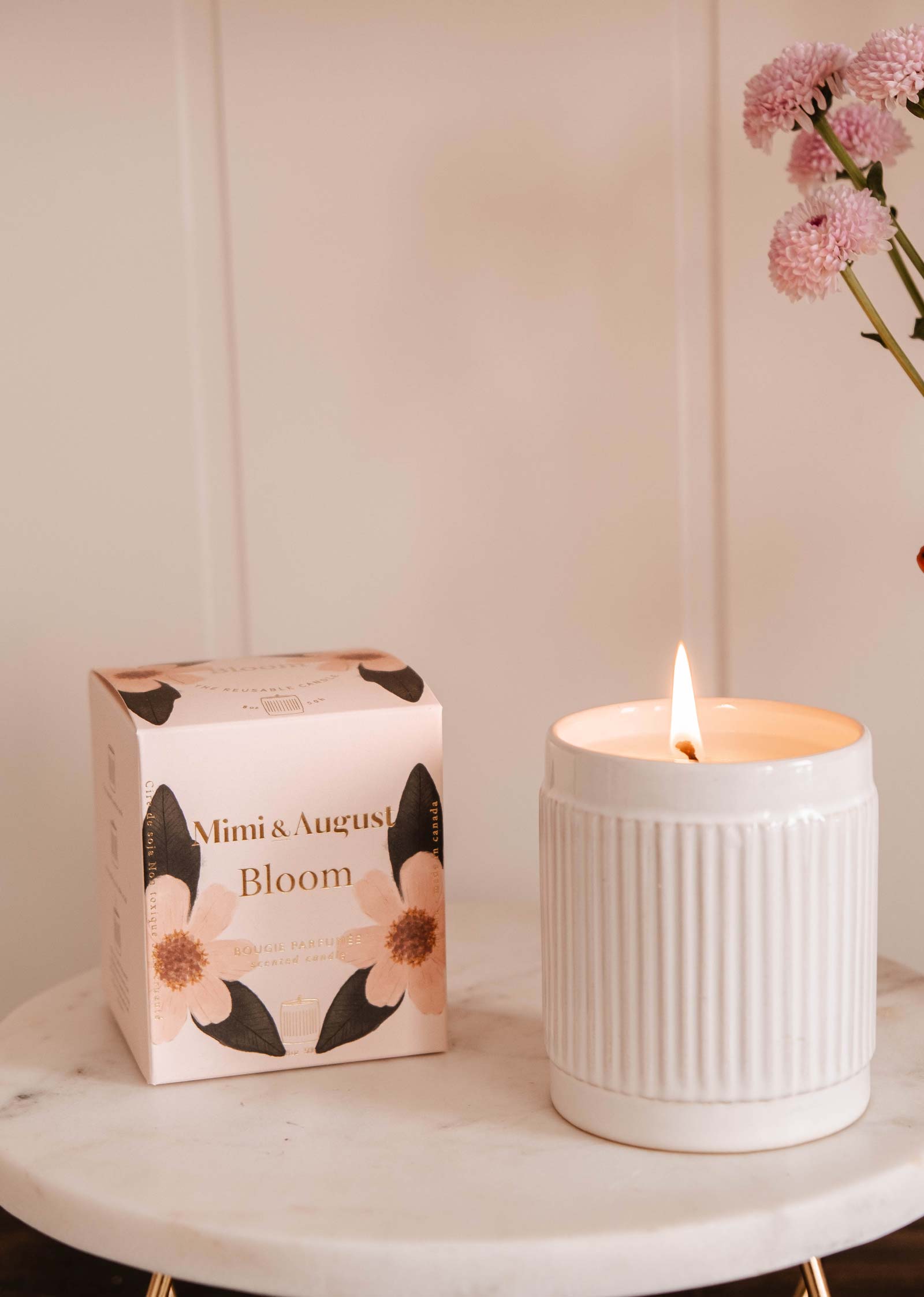 A fruity-scented candle in a white ceramic holder casts a warm glow on the table next to the floral-designed "Mimi & August Bloom - Reusable Candle" box, surrounded by pink flowers.