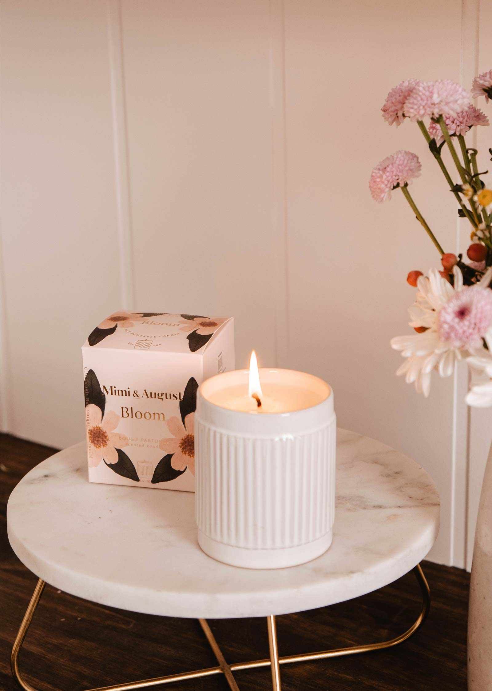 The Bloom – Reusable Soy Candle by Mimi & August, in a ribbed holder, glows on a round marble table beside a floral box and pink flowers in a vase.
