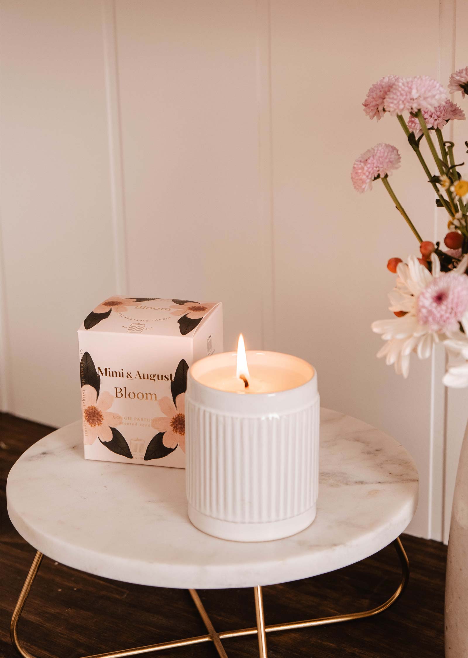 A lit Mimi & August Bloom - Reusable Candle in a white ceramic holder sits on a round marble table, exuding a fruity scent beside a flower bouquet and its box.