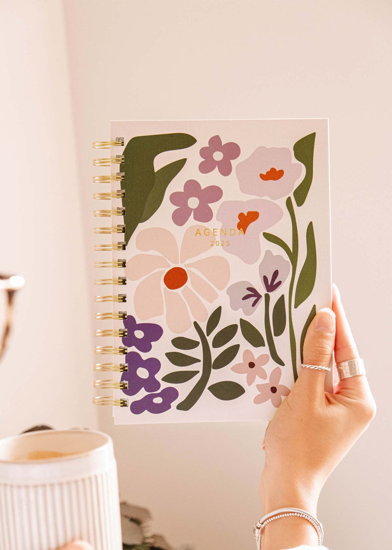 Une main tient le Blooming - Agenda 2025 de Mimi & August, dont la couverture est joliment illustrée à la main d'un motif floral, parfaitement assorti à une tasse de café et idéal pour l'organisation de la journée.