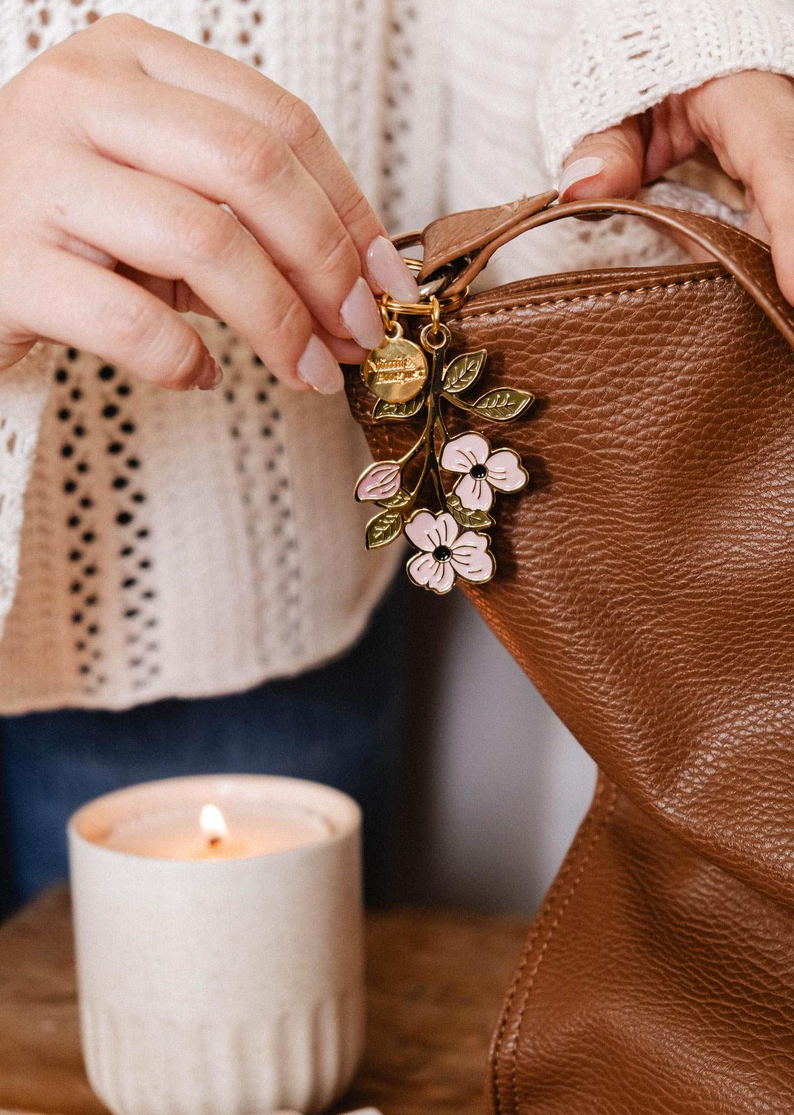 Someone adds the Mimi & August Blossom Branch Keychain to a brown leather bag, with a lit candle nearby on a wooden surface.