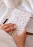 A person holds the Blossom Notebook by Mimi & August, featuring a floral cover labeled "NOTES," with a pen nearby on a white cloth surface.
