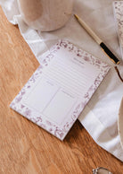 The Blossom - Notepad by Mimi & August, crafted from recycled paper with a floral design, sits on a wooden table next to a pen, white cloth, and ceramic pieces.