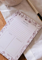 The Blossom - Notepad by Mimi & August, made from recycled paper and featuring sections for notes and important items, is displayed on a cloth surface next to a pen.