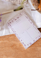 The Blossom - Notepad by Mimi & August, made from recycled paper with sections for to-do lists, notes, and important tasks, rests on a wooden table next to a pen and a plate of fruit.