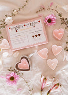 A set of heart-shaped soy wax candles from Mimi & August, housed in a Blush Hearts Candle Tealight box with romantic floral designs, is surrounded by pink flowers on a fabric surface.