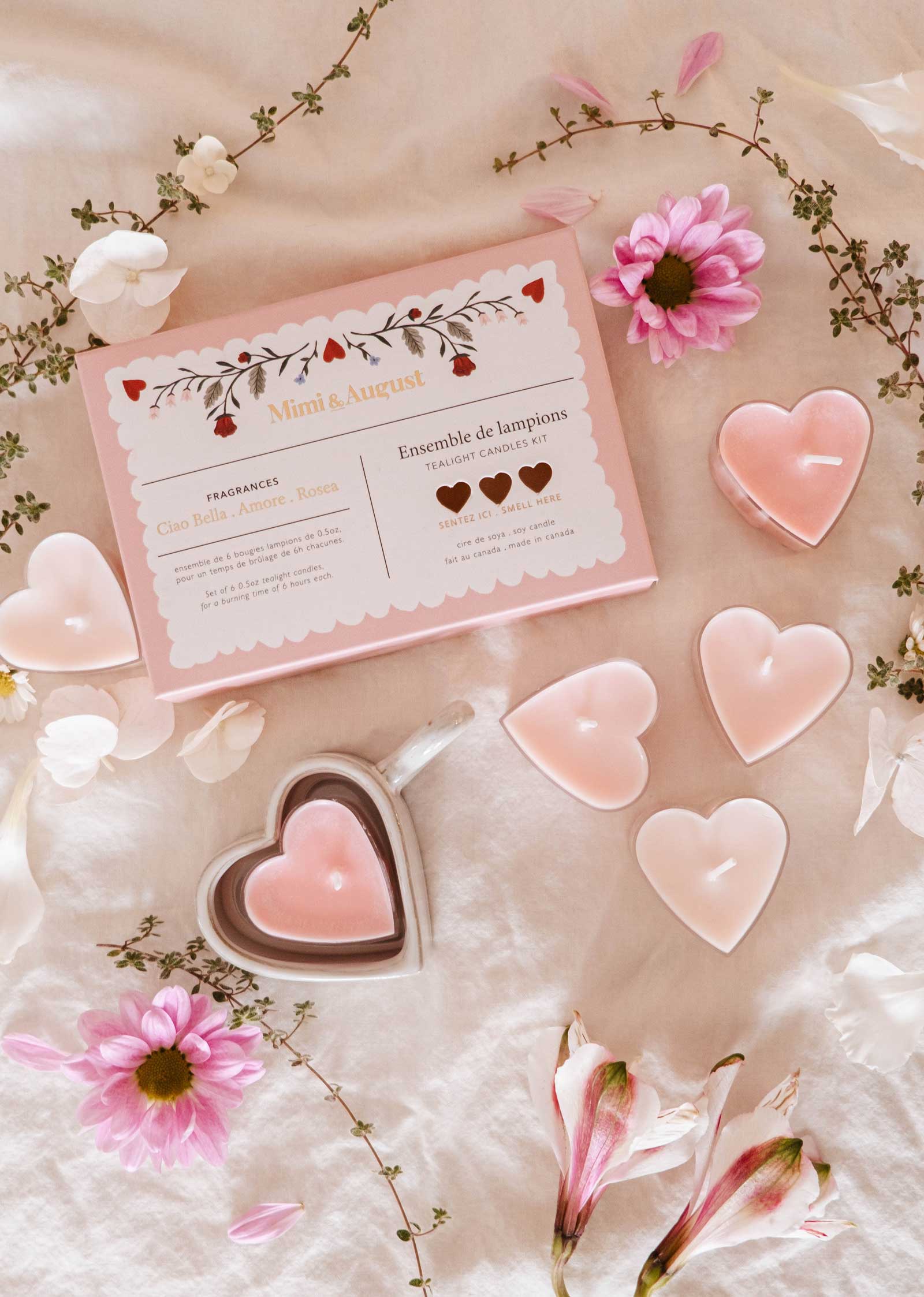 A set of heart-shaped soy wax candles from Mimi & August, housed in a Blush Hearts Candle Tealight box with romantic floral designs, is surrounded by pink flowers on a fabric surface.