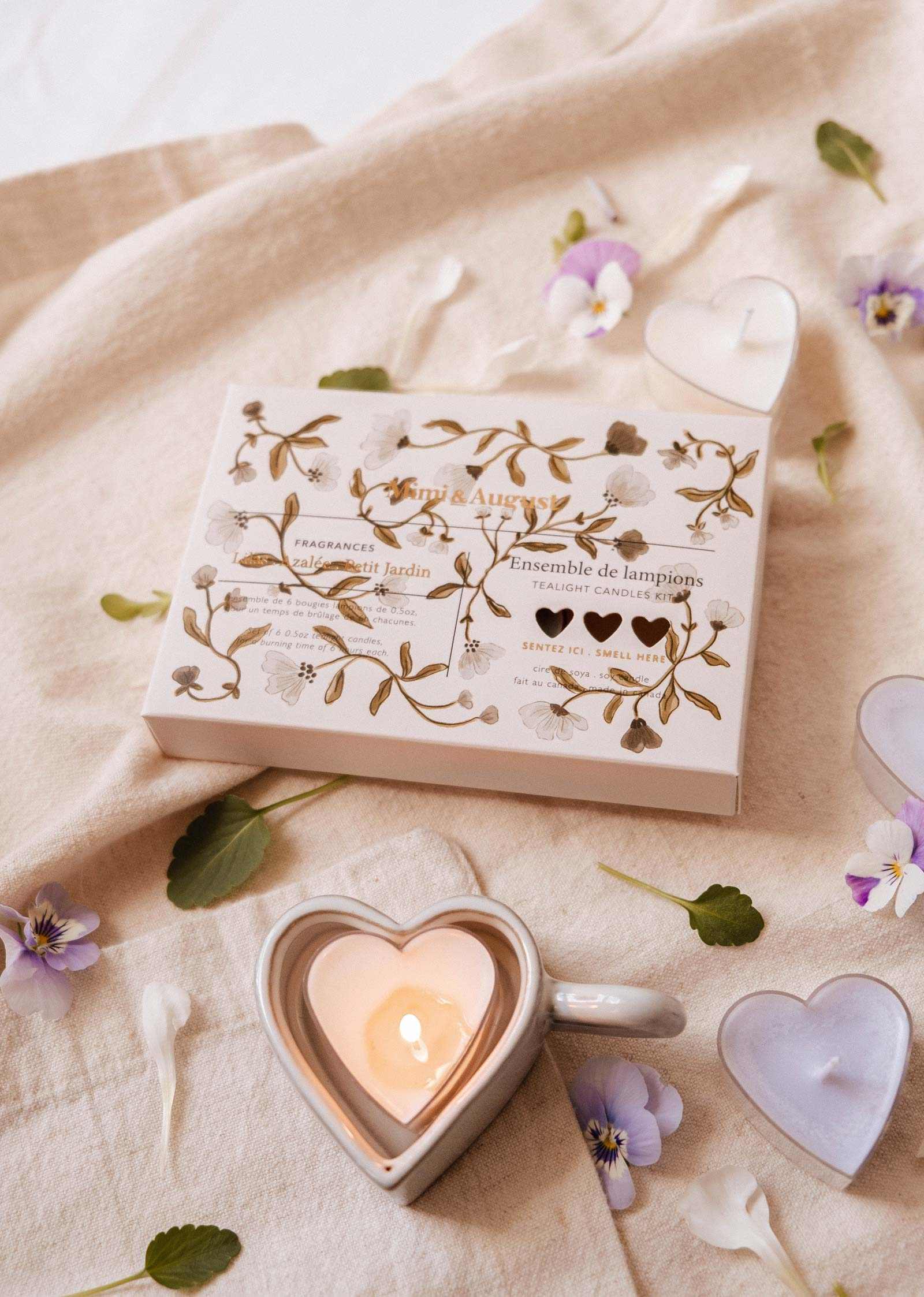 A heart-shaped soy candle from the Mimi & August Blush Hearts Tealight Candle Kit sits in a mug on beige fabric, near its decorative set box and scattered flower petals.