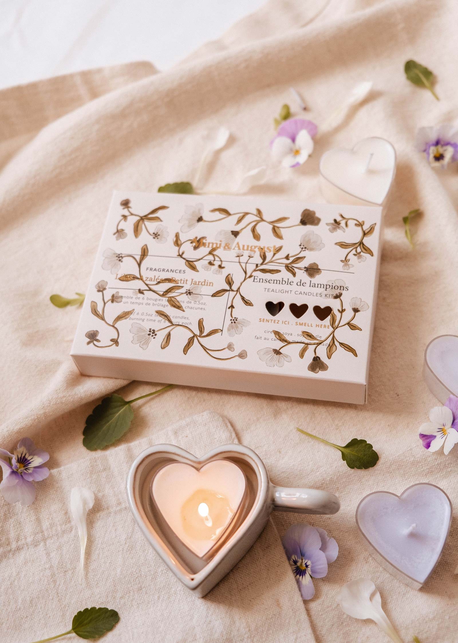 Une boîte de bougies à réchaud Blush Hearts de Mimi & August est posée sur un tissu beige à côté d'une tasse avec une bougie en forme de cœur allumée, entourée de pétales violets et de petites bougies en forme de cœur, créant ainsi une ambiance romantique.