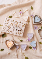 La bougie à réchaud Blush Hearts Candle de Mimi & August présente des bougies en cire de soja pastel en forme de cœur, joliment disposées sur un tissu beige avec des pétales de fleurs et des feuilles.