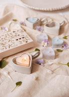Les bougies à réchaud Blush Hearts de Mimi & August, fabriquées à partir de cire de soja, sont en forme de cœur et présentées sur un tissu doux avec des pétales de fleurs éparses et une boîte décorative à proximité.