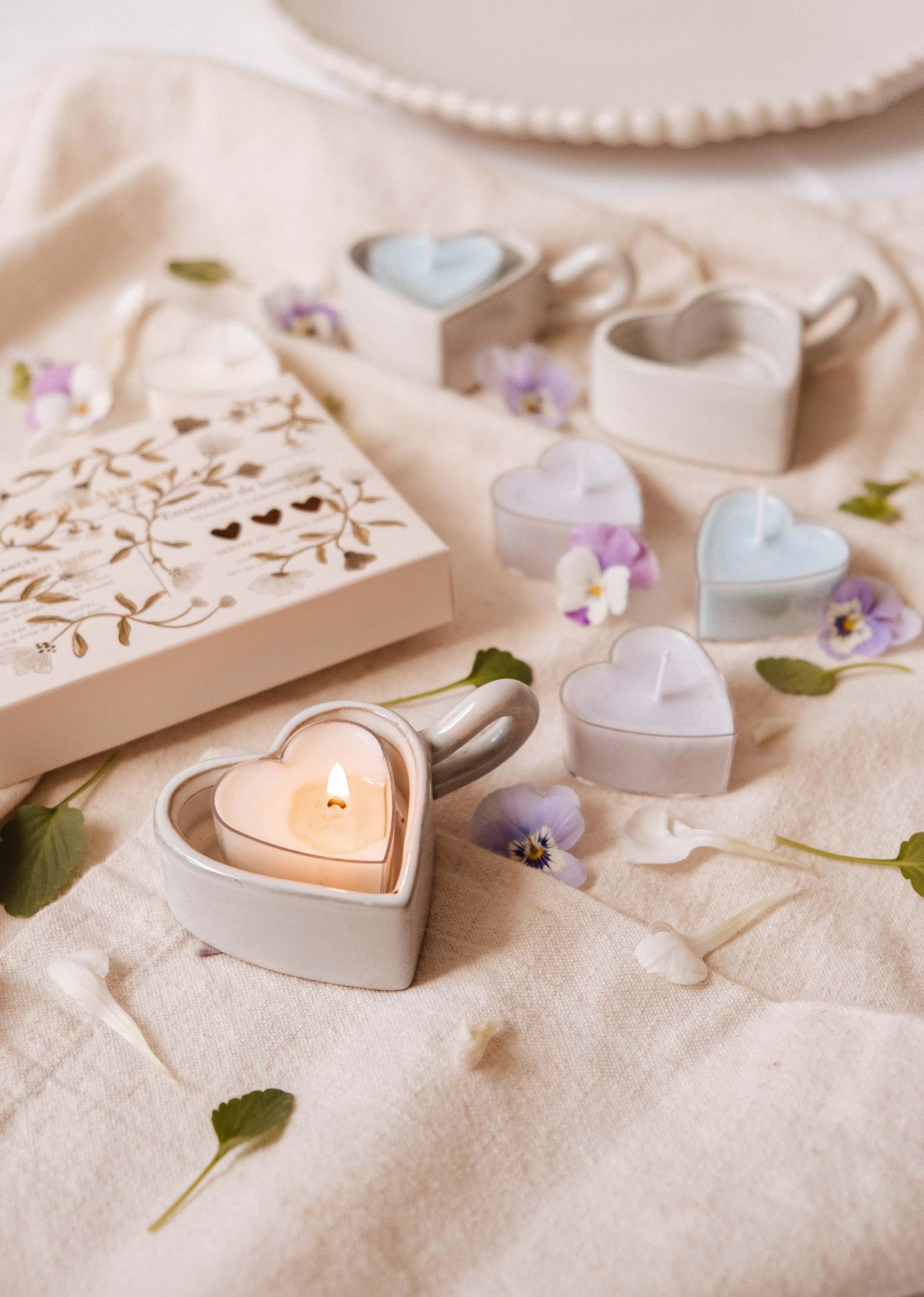 Les bougies à réchaud Blush Hearts de Mimi & August, fabriquées à partir de cire de soja, sont en forme de cœur et présentées sur un tissu doux avec des pétales de fleurs éparses et une boîte décorative à proximité.