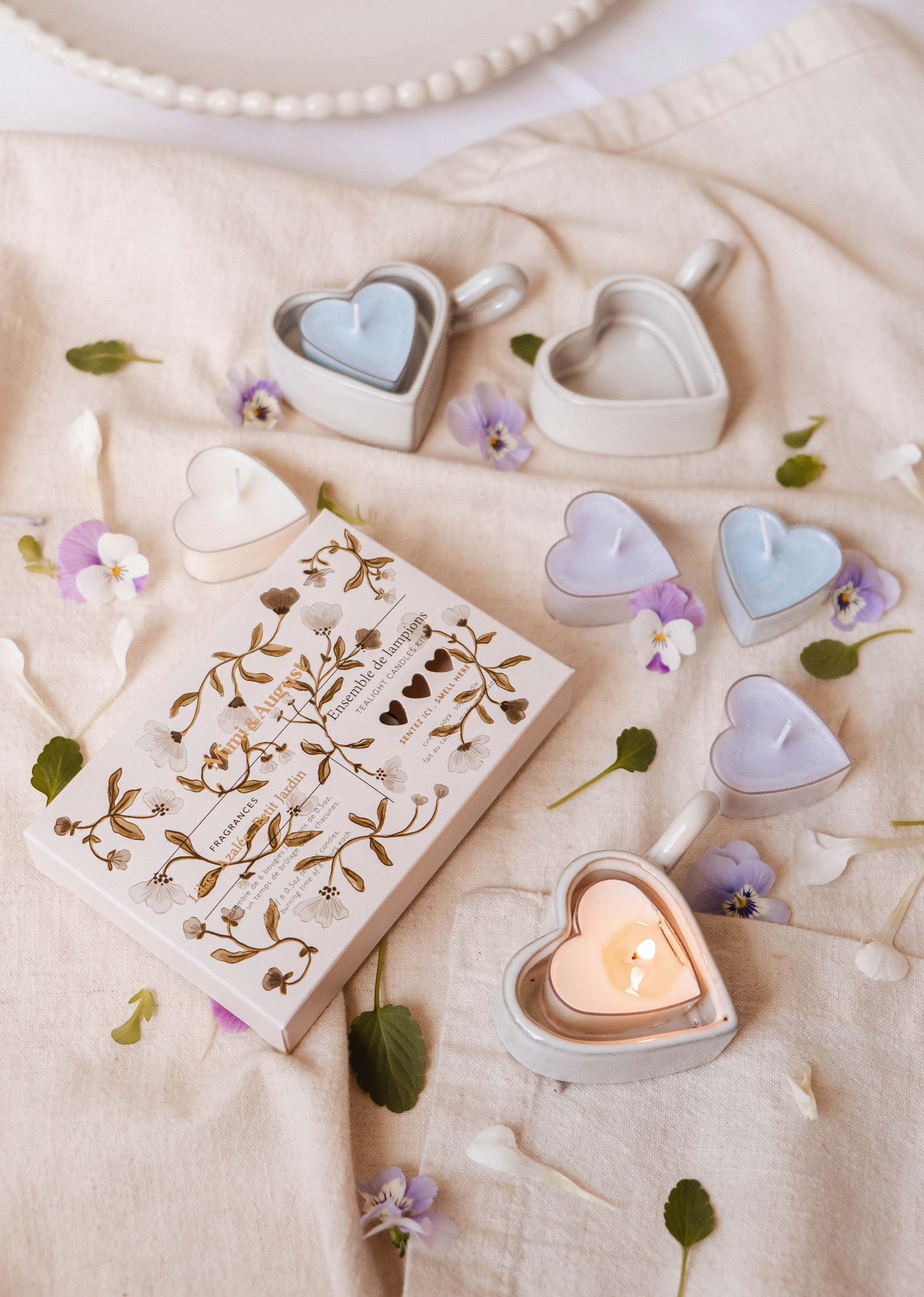 Une mise en page plate présente les bougies Blush Hearts Candle Tealights de Mimi & August dans des plats en céramique en forme de cœur, un carnet à motifs floraux et des petites fleurs éparpillées sur un tissu beige.