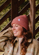 A person wearing a wool coat and a Blush Pink Super Puff Beanie by Mimi & August leans against a wooden structure, smiling.