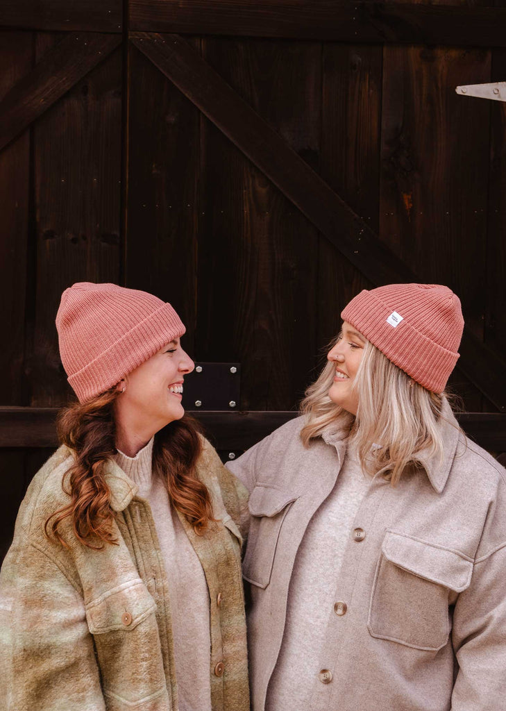 Deux personnes portant un bonnet Blush Pink Super Puff Beanie de Mimi & August et des vestes de couleur claire se tiennent côte à côte, se souriant l'une à l'autre devant une toile de fond en bois - des compléments parfaits à toute garde-robe d'hiver.