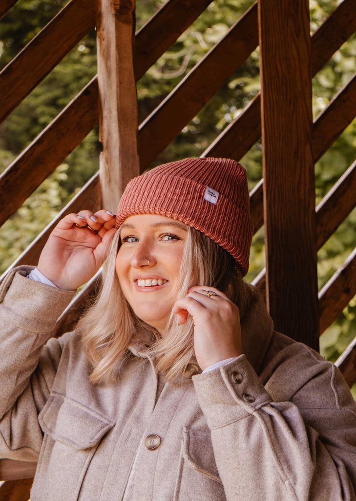 Une personne portant un manteau beige et le bonnet Blush Pink Super Puff Beanie de Mimi & August sourit, tenant les côtés du bonnet, debout devant une balustrade en bois, ajoutant du charme à sa garde-robe d'hiver.