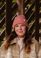 A woman stands outdoors wearing a Blush Pink Super Puff Beanie by Mimi & August and a light-colored jacket, with a wooden structure and foliage in the background.