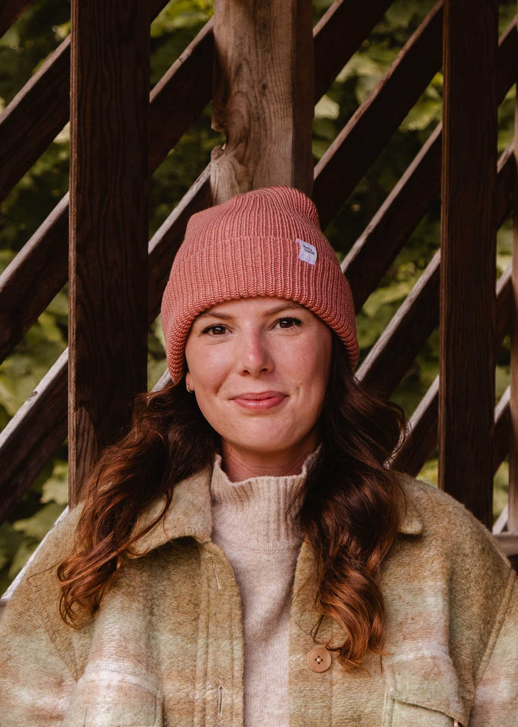 Une femme se tient à l'extérieur, vêtue d'un bonnet Blush Pink Super Puff Beanie de Mimi & August et d'une veste de couleur claire, avec une structure en bois et du feuillage en arrière-plan.