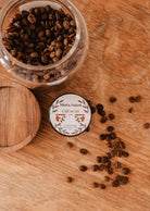 A glass jar of coffee beans sits on wood beside the Mimi & August Café au lait – 2 oz Soy Candle, coffee scented and hand-poured in Montreal, with scattered beans nearby.
