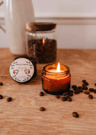 The Café au lait – 2 oz Soy Candle by Mimi & August sits in a brown glass jar on a wooden surface with coffee beans, featuring a reusable, hand-poured soy wax blend made in Montreal. A jar of beans and candle lid are visible in the background.