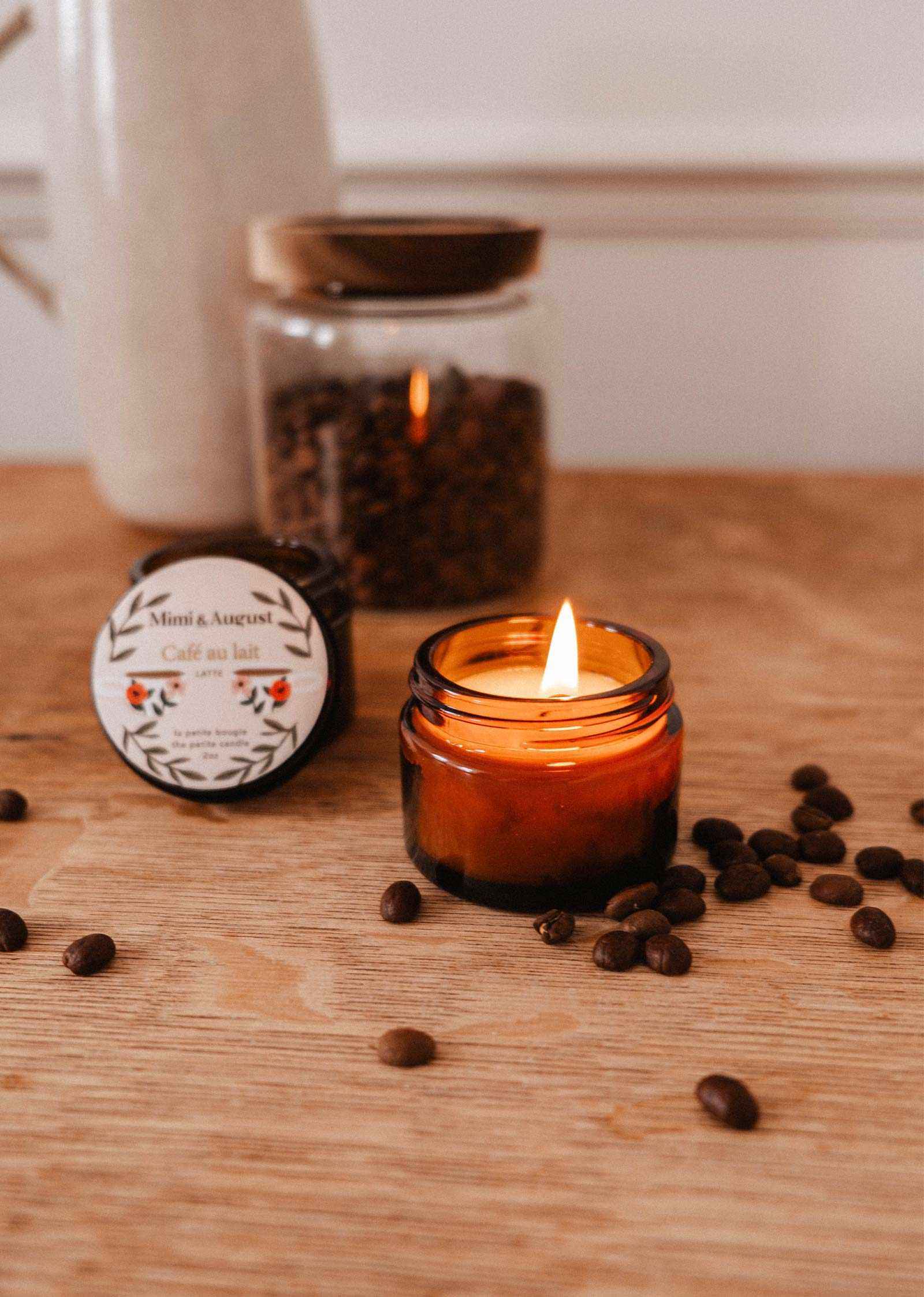 The Café au lait – 2 oz Soy Candle by Mimi & August sits in a brown glass jar on a wooden surface with coffee beans, featuring a reusable, hand-poured soy wax blend made in Montreal. A jar of beans and candle lid are visible in the background.