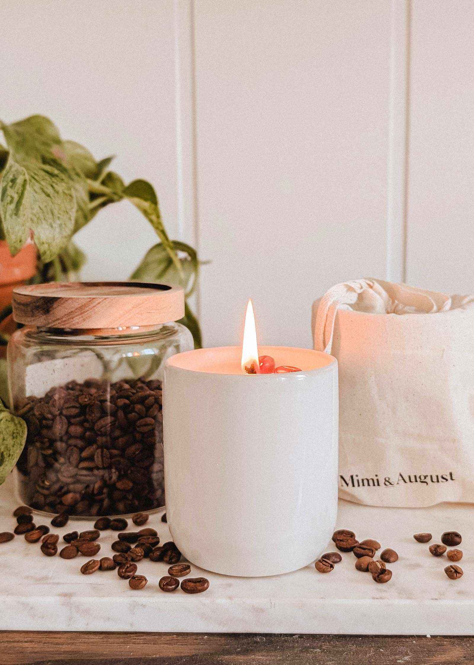 A lit Mimi & August Special Edition Candle in a white holder rests on marble beside a jar of coffee beans, a cloth bag, and a potted plant, with scattered coffee beans completing the scene.